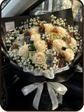 A product listing graphic for a 'Grand Celebration Mix' bouquet from DrapedNuts. The image features a photo of a black flower box containing a mix of white and black roses, chocolates, and biscuits, with a silver ribbon.
